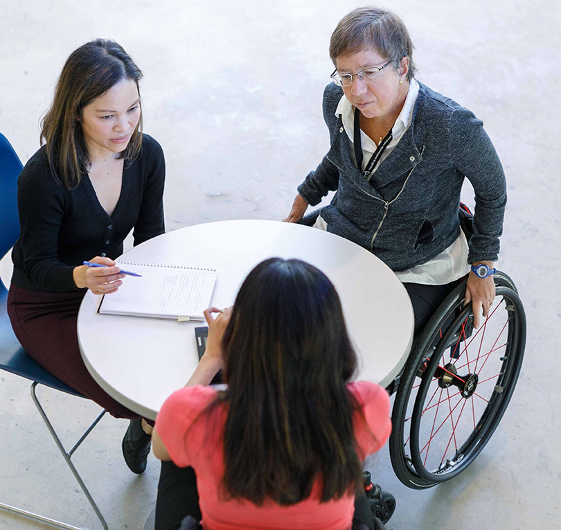 Three people, including two in wheelchairs, sit at a table discussing a document